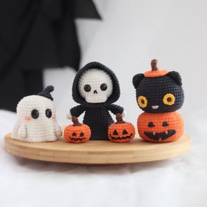 May include: A collection of handmade crochet Halloween decorations. Includes a white ghost, a skeleton in a black hooded robe, and a black cat on top of an orange pumpkin. The decorations are displayed on a round wooden tray.