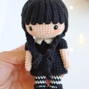 May include: A handmade crochet doll with black hair in braids, wearing a black dress with a white collar, and black and white striped socks. The doll has a light skin tone and rosy cheeks. The doll is approximately 5 inches tall.