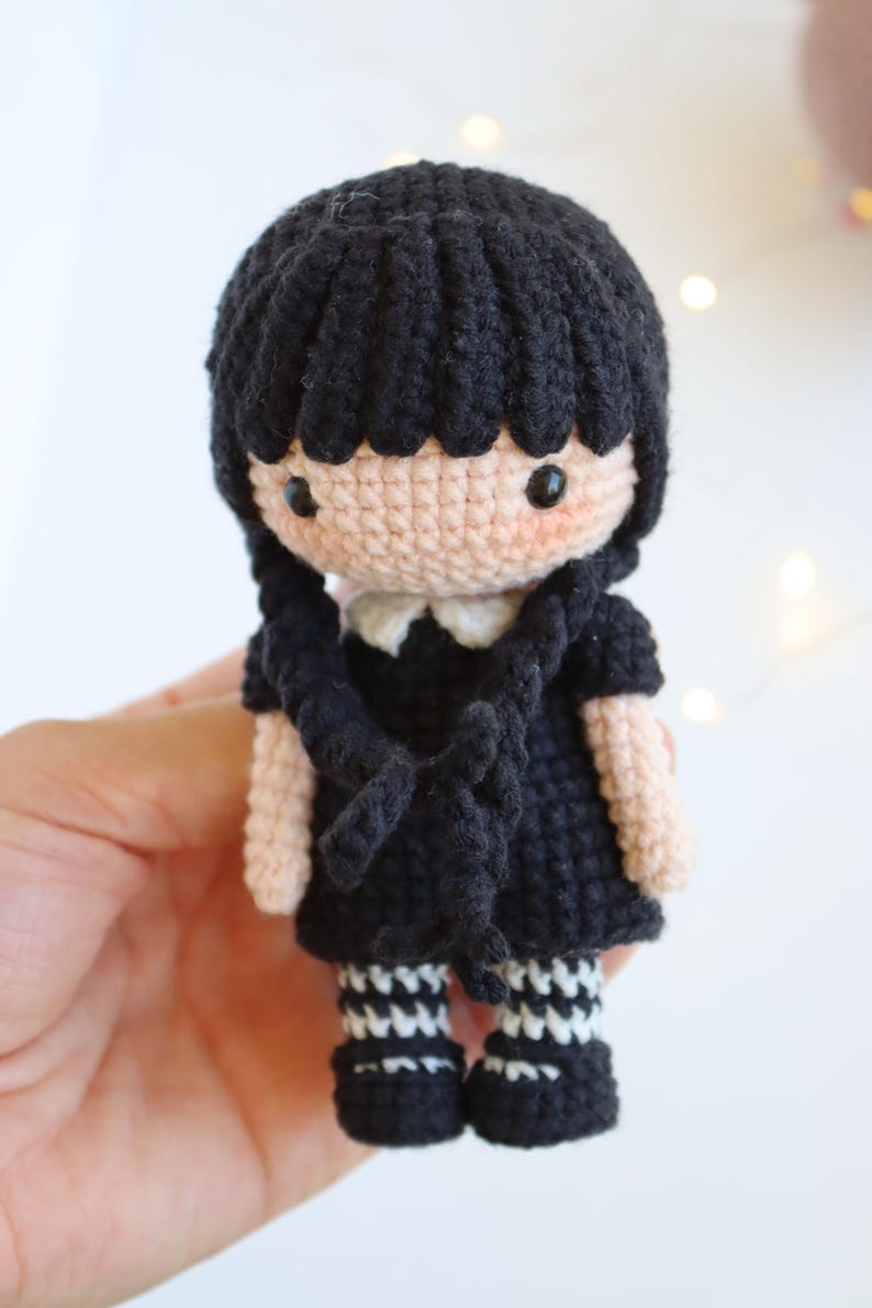 May include: A handmade crochet doll with black hair in braids, wearing a black dress with a white collar, and black and white striped socks. The doll has a light skin tone and rosy cheeks. The doll is approximately 5 inches tall.