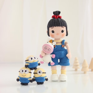 Könnte beinhalten: Gehäkelte Puppe in blauen Latzhosen, die ein Einhorn-Plüschtier hält, mit drei gehäkelten Minion-Figuren. Die Puppe hat schwarzes Haar und eine rote Haarschleife. Die Minions sind gelb mit blauen Latzhosen und Schutzbrillen. Der Hintergrund ist weiß.