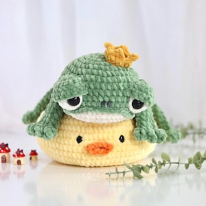Può includere: Un peluche a forma di rana lavorato all'uncinetto di colore verde con una piccola corona gialla sulla testa, appoggiato sopra un peluche a forma di papera rotonda lavorato all'uncinetto di colore giallo. La rana ha grandi occhi neri e pancia bianca.