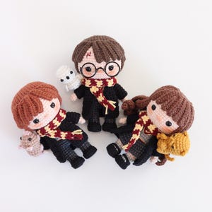May include: Three crocheted dolls depicting characters from a well-known fantasy series. Each doll has a distinctive hairstyle, glasses, and a scarf in red, gold, and black. One doll holds a white owl, and another holds a brown mouse.