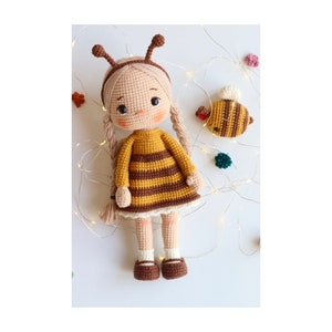 May include: A crocheted doll dressed as a bee with brown and yellow stripes. The doll has blonde hair and is wearing a white dress underneath the bee costume. The doll is holding a small crocheted bee.