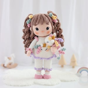 以下が含まれることがあります： 茶色の巻き毛をツインテールにした手作りの編みぐるみの人形。紫色の縁取りがあるクリーム色の服を着ています。人形は小さな白いユニコーンのぬいぐるみを抱えており、髪と服にはパステルカラーのアクセントがあしらわれています。