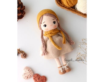 May include: A crocheted doll with brown hair, a yellow hat, a yellow scarf, and a beige dress. The doll is wearing brown shoes and is standing on a white surface. The doll is surrounded by hearts and other crocheted items.