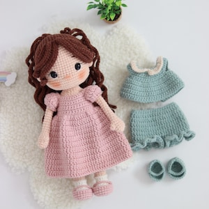 May include: A handmade crochet doll with brown yarn hair, wearing a pink dress and matching shoes. Included is an additional set of clothing consisting of a sage green top with a cream-colored collar and matching ruffled shorts with two small shoes.