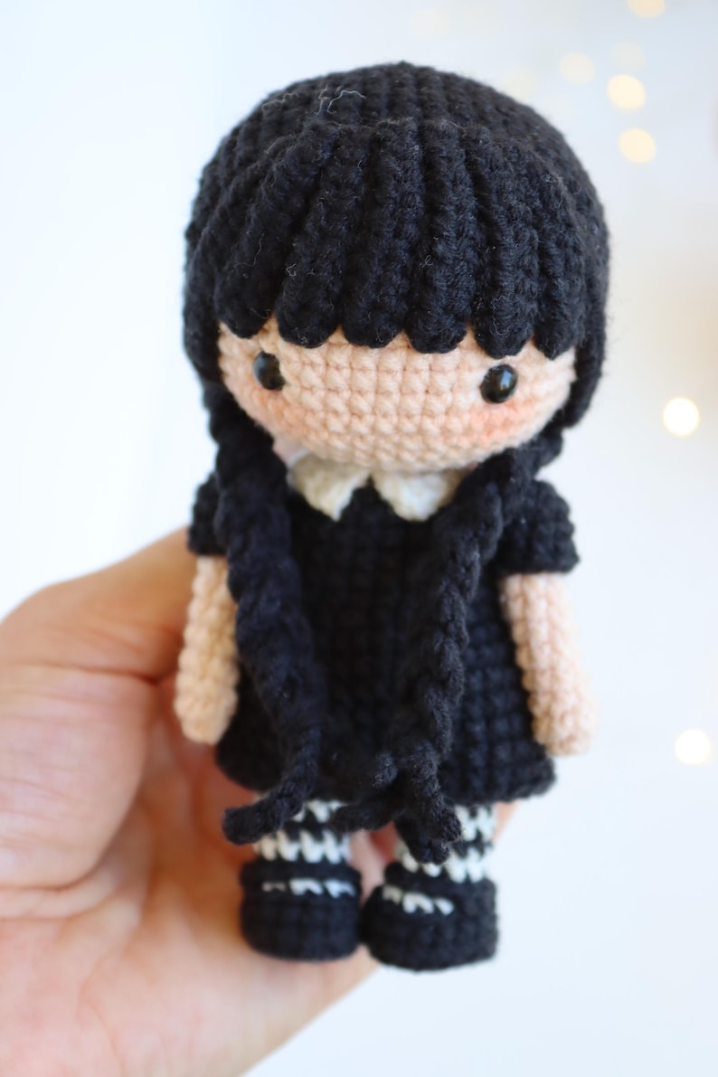 May include: Crocheted doll with black hair, a black dress, and white collar. The doll has black eyes, rosy cheeks, and black and white striped stockings. The doll is a handmade craft item.