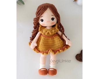 Peut inclure: Une poupée en crochet aux longs cheveux bruns, portant une robe jaune avec des bordures brunes et des chaussures brunes. La poupée est debout sur une surface blanche.