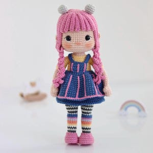 Peut inclure: Une poupée au crochet faite à la main avec des cheveux roses tressés et deux petits accents en forme de boule. La poupée porte une robe bleue avec une bordure rose, des leggings rayés et des chaussures roses. Un article charmant et artisanal.