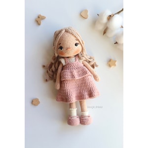 May include: A crocheted doll with long blonde hair, wearing a pink dress and white socks. The doll is standing on a white surface with wooden decorations.