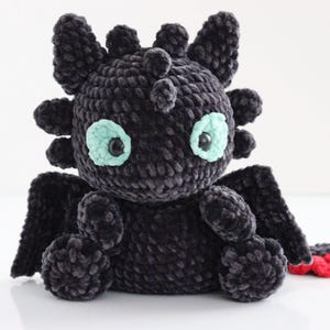 May include: A handmade black crochet dragon plushie with mint green eyes and small wings. The dragon has a round body, small legs, and a tail with red accents. The plushie is made of soft, textured yarn.