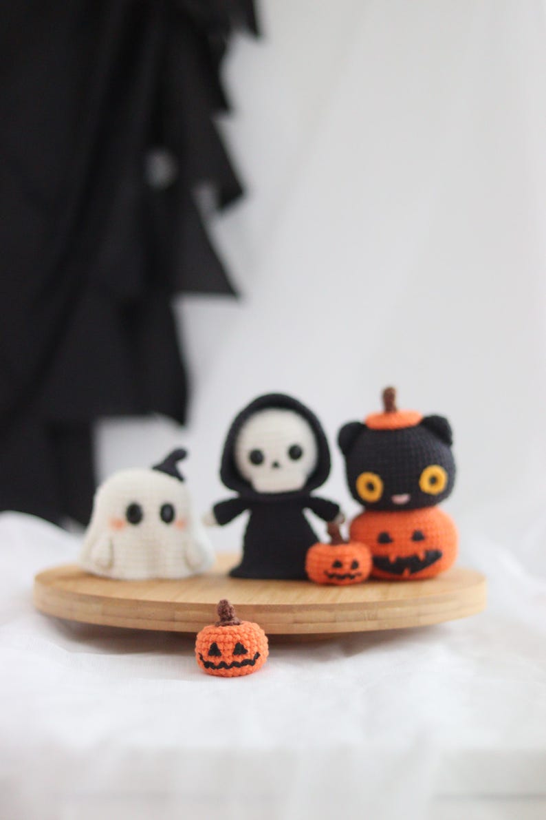 May include: A collection of handmade crochet Halloween decorations. Includes a white ghost, a black Grim Reaper, a black cat with an orange pumpkin body, and two orange pumpkins. The figures are displayed on a wooden tray.
