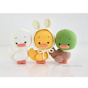 May include: Three handmade crochet ducklings in white, yellow, and green. The yellow duckling wears a bunny hat. Each duckling has a peach-coloured beak and feet. The ducklings are displayed on a white background.