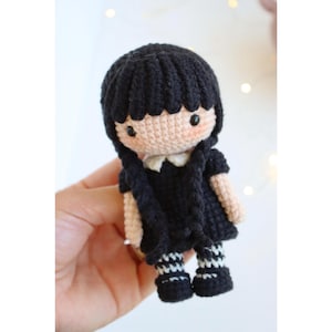 Puede incluir: Muñeca de ganchillo hecha a mano con cabello negro, un vestido negro y calcetines a rayas. La muñeca tiene un tono de piel claro, mejillas rosadas y un cuello blanco. El cabello de la muñeca está peinado en dos trenzas.