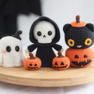 May include: A collection of handmade crochet Halloween decorations. Includes a white ghost, a skeleton in a black robe, two orange pumpkins, and a black cat on top of a pumpkin. All are displayed on a round wooden tray.