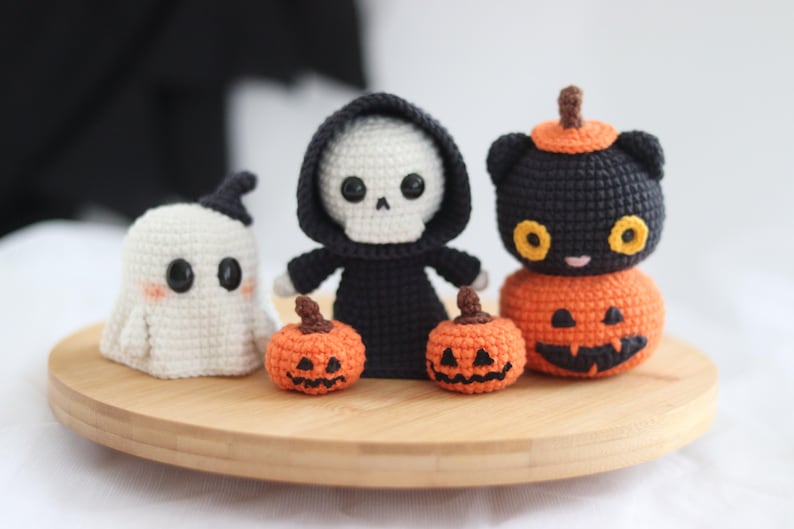 May include: A collection of handmade crochet Halloween decorations. Includes a white ghost, a skeleton in a black robe, two orange pumpkins, and a black cat on top of a pumpkin. All are displayed on a round wooden tray.