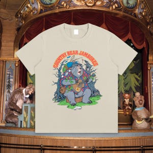 Könnte beinhalten: Ein cremefarbenes T-Shirt mit einem Cartoon-Bären, der Ukulele spielt. Der Bär trägt ein kariertes Hemd und einen Hut, mit den Worten "COUNTRY BEAR JAMBOREE" in einem geschwungenen Design darüber. Das T-Shirt hat einen klassischen Rundhalsausschnitt.