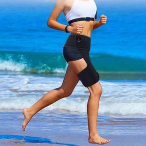 May include: A person running on a beach wearing a black thigh support brace. The brace is worn on the left leg and is secured with a velcro strap. The person is also wearing black shorts and a white sports bra.