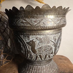 May include: A dark metal planter with a scalloped rim and a wide, flared body. The surface features intricate etched designs of deer and floral motifs, with a textured, darkened finish that highlights the engraved patterns throughout the piece.