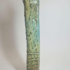 May include: A tall, textured ceramic vase with a flared top. The vase has a cylindrical body with a rough, organic texture and a gradient of blue and green hues. The base is a darker, earthy tone.