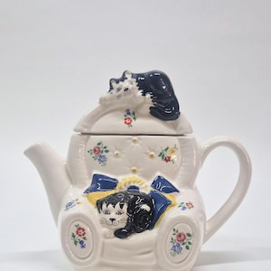 May include: A white ceramic teapot with a cat design. The teapot has a black and white cat on the lid and another on the front, resting on a cushion. Floral and bow details decorate the teapot.