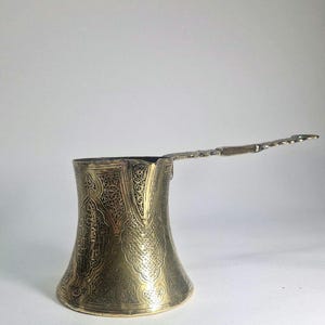 May include: A brass-coloured Turkish coffee pot with an ornate, engraved design. The pot has a flared base, a narrow neck, and a long, flat handle. The handle is also decorated with an engraved pattern.