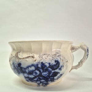 May include: A cream-coloured ceramic chamber pot featuring a deep blue floral pattern and intricate embossed scrollwork details. The vessel has a wide, fluted rim, a rounded body, and a single ornate handle extending from the side.