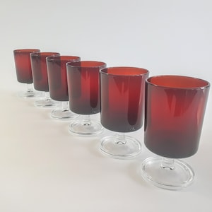 May include: Six clear glass wine glasses with a deep red tint. The glasses are arranged in a row from left to right, with the tallest glass on the left and the shortest glass on the right.