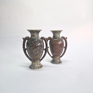 May include: Two small, brown and green ceramic vases with handles. The vases have a speckled glaze and a textured surface.