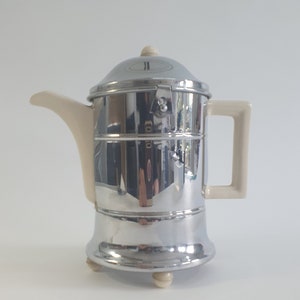 May include: A chrome and white ceramic teapot with a lid and a handle. The teapot has a round base with three feet.