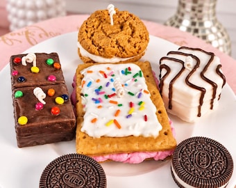 May include: A collection of food-shaped candles on a white plate, including a chocolate brownie with colorful candies, a cookie sandwich, a rectangular pastry with white frosting and sprinkles, a white frosted cake, and two round Oreo cookies.