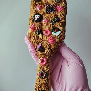 May include: A handheld mirror with a handle and frame covered in textured tan whipped cream-like material. The surface is decorated with miniature charms including pink ice cream cones, chocolate donuts, and small cakes with colorful sprinkles.