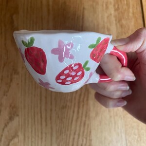 May include: A white ceramic mug with a pink handle. The mug is decorated with hand-painted pink flowers and red strawberries.