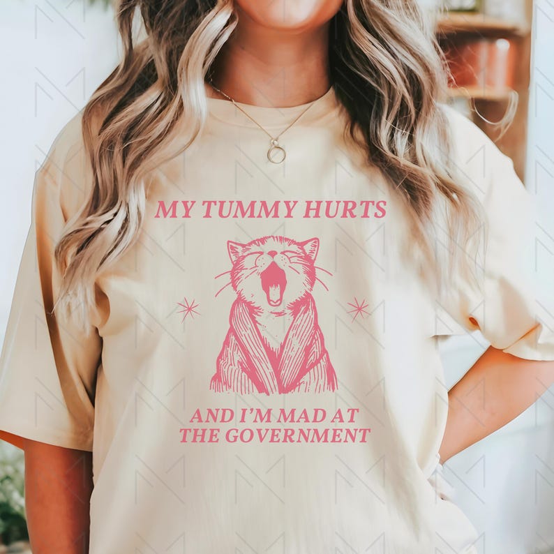 May include: A light yellow t-shirt with a pink graphic of a cat with its mouth open. The text "My tummy hurts and I'm mad at the government" is printed on the shirt.