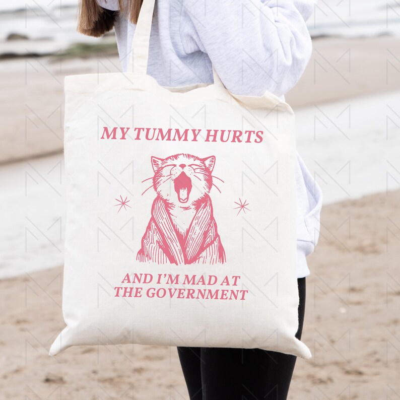May include: A white tote bag with a pink cat illustration and the text "My tummy hurts and I'm mad at the government".
