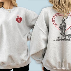May include: Pale grey sweatshirt featuring a red heart-shaped design on the front and back. The front has a heart-shaped locket with a cherub. The back displays a knight within a heart, with the text "OH BARTHOLOMEW LIFE IS DREAMY".