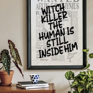 May include: A framed print with a vintage newspaper background. The text "WANTED" is at the top of the print. The text "WITCH KILLER THE HUMAN IS STILL INSIDE HIM" is written in black paint over the newspaper text.