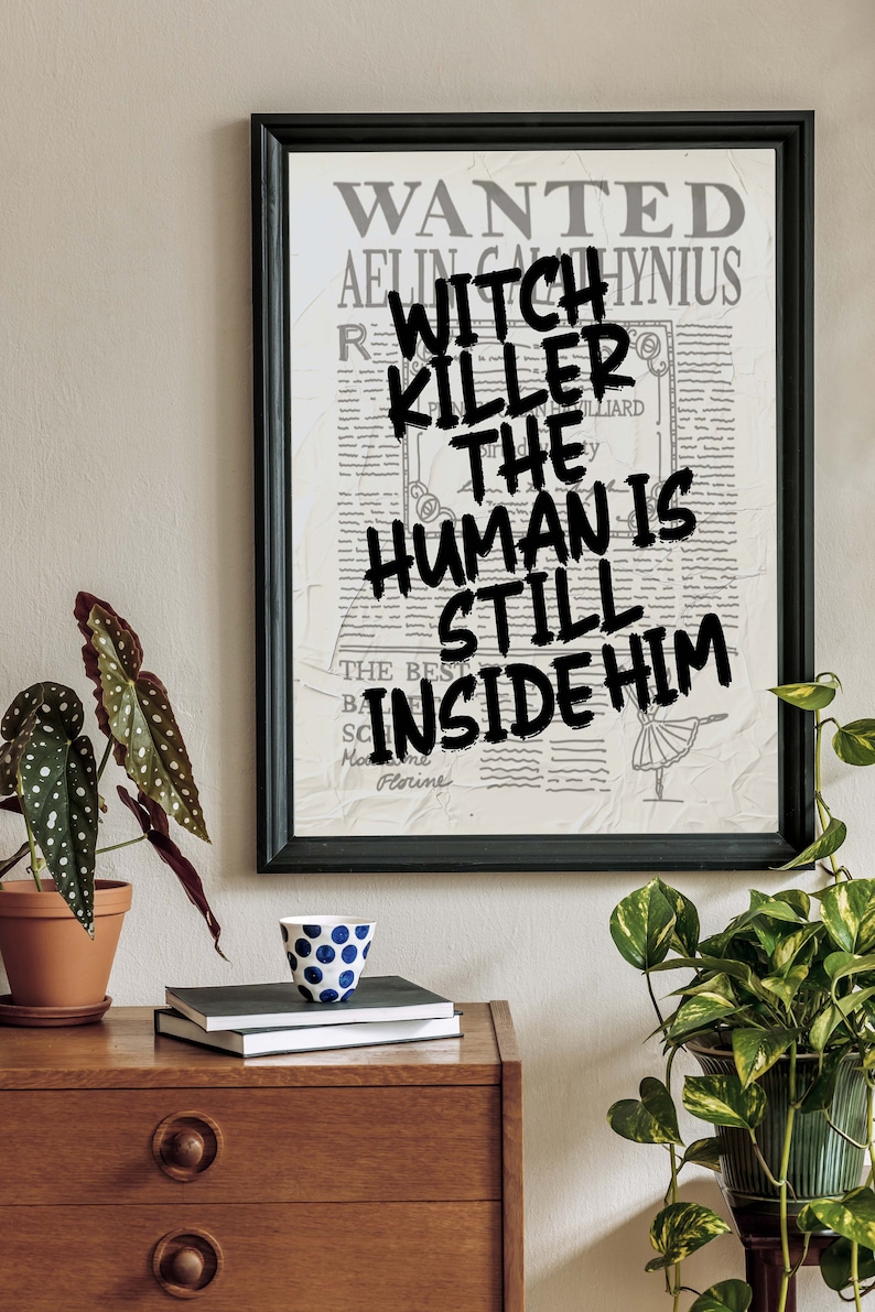 May include: A framed print with a vintage newspaper background. The text "WANTED" is at the top of the print. The text "WITCH KILLER THE HUMAN IS STILL INSIDE HIM" is written in black paint over the newspaper text.
