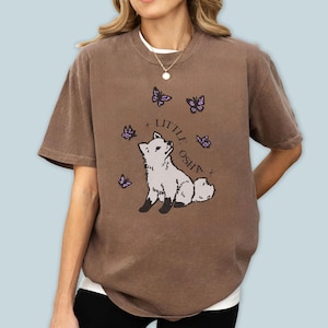 Può includere: T-shirt marrone con un disegno di volpe dei cartoni animati, la scritta "LITTLE OSH" e farfalle viola. La maglietta ha una vestibilità ampia e maniche corte. La volpe è grigia e bianca, con dettagli neri.