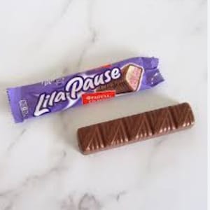 Pode incluir: Uma barra de chocolate Lila Pause e uma barra parcialmente desembrulhada sobre uma superfície de mármore branco. A embalagem é roxa com o texto "Lila Pause" em escrita branca. A barra de chocolate é marrom e tem um design canelado.