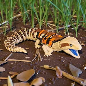 May include: A detailed sculpture of a lizard with a cream-coloured head and a body featuring brown, black, and orange stripes. The lizard's mouth is open, revealing a blue tongue. It rests on a bed of soil with small twigs and blades of grass.