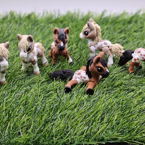 May include: A collection of seven small, colourful, plastic toy horses. The horses are in various poses, including standing, lying down, and running. The horses are all different colours, including brown, white, black, and tan. The horses are on a green grassy surface.