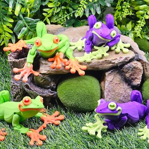 May include: Four articulated frog figurines, two green and two purple, posed on a rock and moss covered surface. The frogs have bright orange feet and eyes.