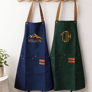 Peut inclure: Deux tabliers suspendus à un porte-manteau en bois. Le tablier de gauche est bleu marine avec un motif de montagne doré et le texte WILSON. Le tablier de droite est vert foncé avec un monogramme doré TJM. Ils ont des lanières marron et deux poches.