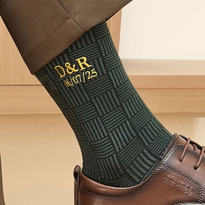 May include: A dark green sock featuring a black woven grid pattern and gold embroidered text that reads D&R followed by the date 06/07/25. The sock is worn with a brown leather lace-up shoe and brown trousers, showcasing a detailed texture.