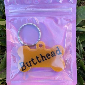 May include: A pink iridescent plastic bag containing a bone-shaped dog tag with the word "Butthead" written in black lettering. The tag is attached to a silver key ring.