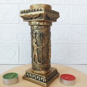 Op de afbeelding: Gouden kaarsenhouder in de vorm van een Egyptische zuil met hiërogliefen en een vrouwenfiguur. De houder staat op een houten oppervlak met twee theelichtjes.