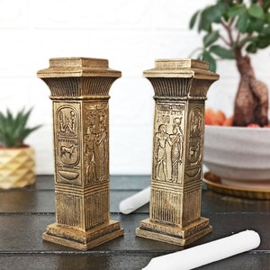 Op de afbeelding: Twee gouden kaarsenstandaards met ingewikkelde Egyptische ontwerpen. De standaards zijn gevormd als kolommen en bevatten hiërogliefen en afbeeldingen van goden en godinnen. Een witte kaars ligt op een donker houten oppervlak.