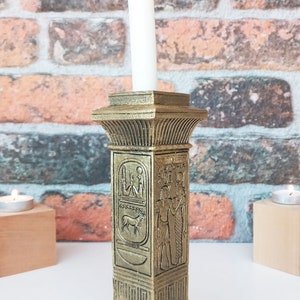 Op de afbeelding: Gouden kleur kaarsenhouder met Egyptische hiërogliefen en een witte kaars. De houder is gevormd als een pilaar met een vierkante basis.