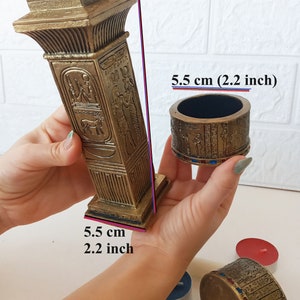 Op de afbeelding: Gouden Egyptische pilaar-kaarshouder met ingewikkelde hiërogliefen-ontwerpen. De kaarshouder is 16 cm hoog en 5,5 cm breed. De basis van de kaarshouder is vierkant en de bovenkant is rond. De kaarshouder is gemaakt van een duurzaam materiaal en is perfect om een ​​toets van Egyptische stijl aan uw huis toe te voegen.